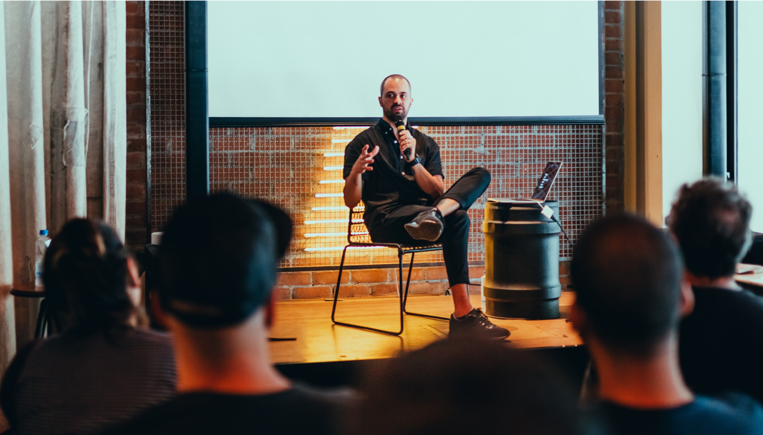 Photograph of a presenter speaking into a microphone seated in a chair on a stage